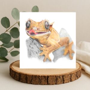 Crested gecko lizard colour pencil drawing. The gecko has its tongue sticking out with big reflected eyes and small eyelashes. The lizard is sat on a rock. The drawing is on a plain white background on a square card. The card is displayed on a log slice with green leaves