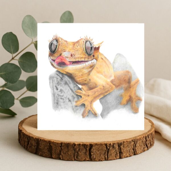 Crested gecko lizard colour pencil drawing. The gecko has its tongue sticking out with big reflected eyes and small eyelashes. The lizard is sat on a rock. The drawing is on a plain white background on a square card. The card is displayed on a log slice with green leaves