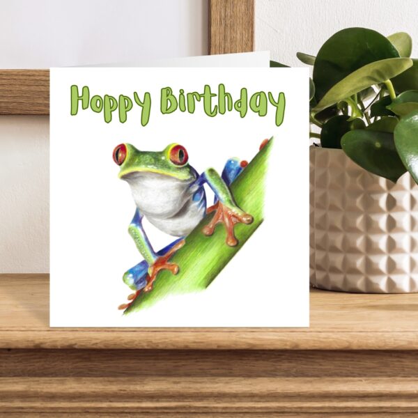 Colour pencil drawing of a Red Eyed Tree Frog on a green stem. The frog is green, orange and blue with big red eyes. The message Hoppy Birthday is written above the frog. The artwork is on a square card displayed on a wooden shelf