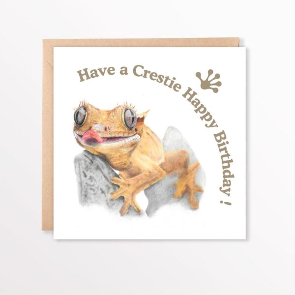 Colour pencil drawing of a crested gecko lizard with large eyes and tongue out. The gecko is clinging to a small rock. There is text that says Have a crestie happy birthday with a gecko foot logo. The art is on the front of a square card on a brown envelope