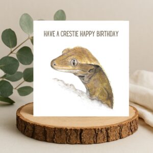 Colour pencil drawing of a crested gecko head and shoulders. A small cute lizard with sweet eyes and eyelashes. There is text that says Have a Crestie Happy Birthday at the top. The artwork is on the front of a square white card sat on a log slice with leaves behind