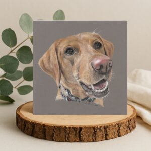 Colour pencil drawing of a Fox Red Labrador Dog. Head and shoulders portrait. The dog has a pink nose and has her mouth open with a happy face. The drawing is against a grey background and is on a square card displayed on a log slice.