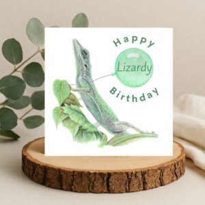 Colour pencil green lizard drawing sitting on some leaves. There is a plain white background and the lizard is holding a green balloon. The text says Happy Lizardy Birthday, with the word Lizardy inside the balloon. The artwork is on a square card sat on a log slice
