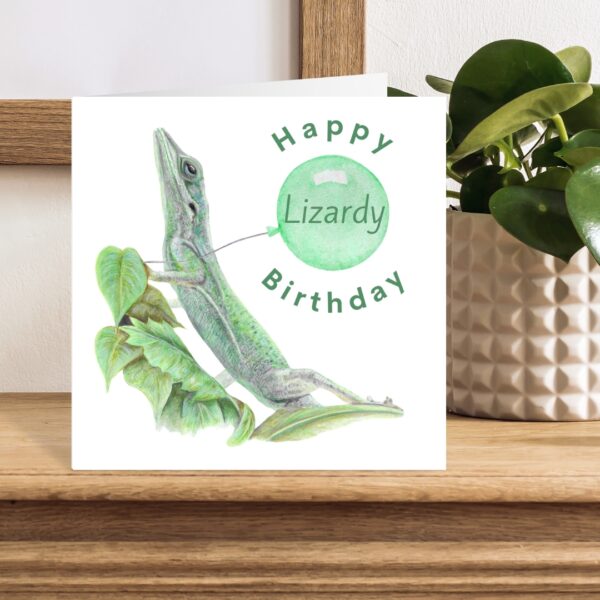 Colour pencil green lizard drawing sitting on some leaves. There is a plain white background and the lizard is holding a green balloon. The text says Happy Lizardy Birthday, with the word Lizardy inside the balloon. The artwork is on a square card sat on a wooden shelf