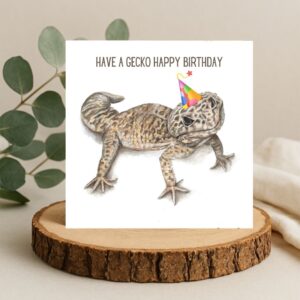 Leopard gecko lizard drawing wearing a party hat. The words Have a gecko happy birthday are written above the gecko. The art is on a plain white background on a square greetings card. The card is displayed on a natural log slice