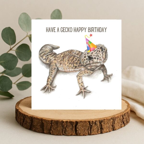 Leopard gecko lizard drawing wearing a party hat. The words Have a gecko happy birthday are written above the gecko. The art is on a plain white background on a square greetings card. The card is displayed on a natural log slice