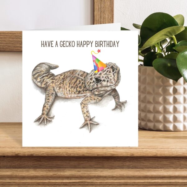 Colour pencil drawing of a leopard gecko lizard wearing a birthday hat against a white background. The art is on the front of a square greetings card