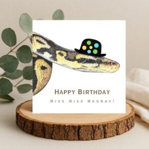 Colour pencil drawing of a yellow and black snake head and shoulders wearing a bowler hat with coloured spots on. The snake is on a white background with the wording Happy Birthday Hiss Hiss Hooray. The card is on a log slice.