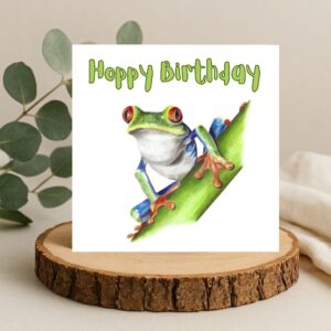 Colour pencil drawing of a Red Eyed Tree Frog on a green stem. The frog is green, orange and blue with big red eyes. The message Hoppy Birthday is written above the frog. The artwork is on a square card displayed on a log slice