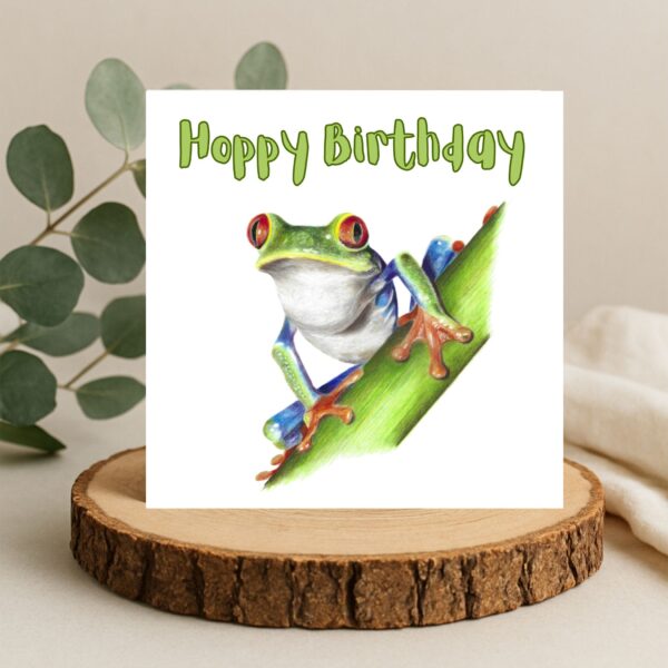Colour pencil drawing of a Red Eyed Tree Frog on a green stem. The frog is green, orange and blue with big red eyes. The message Hoppy Birthday is written above the frog. The artwork is on a square card displayed on a log slice