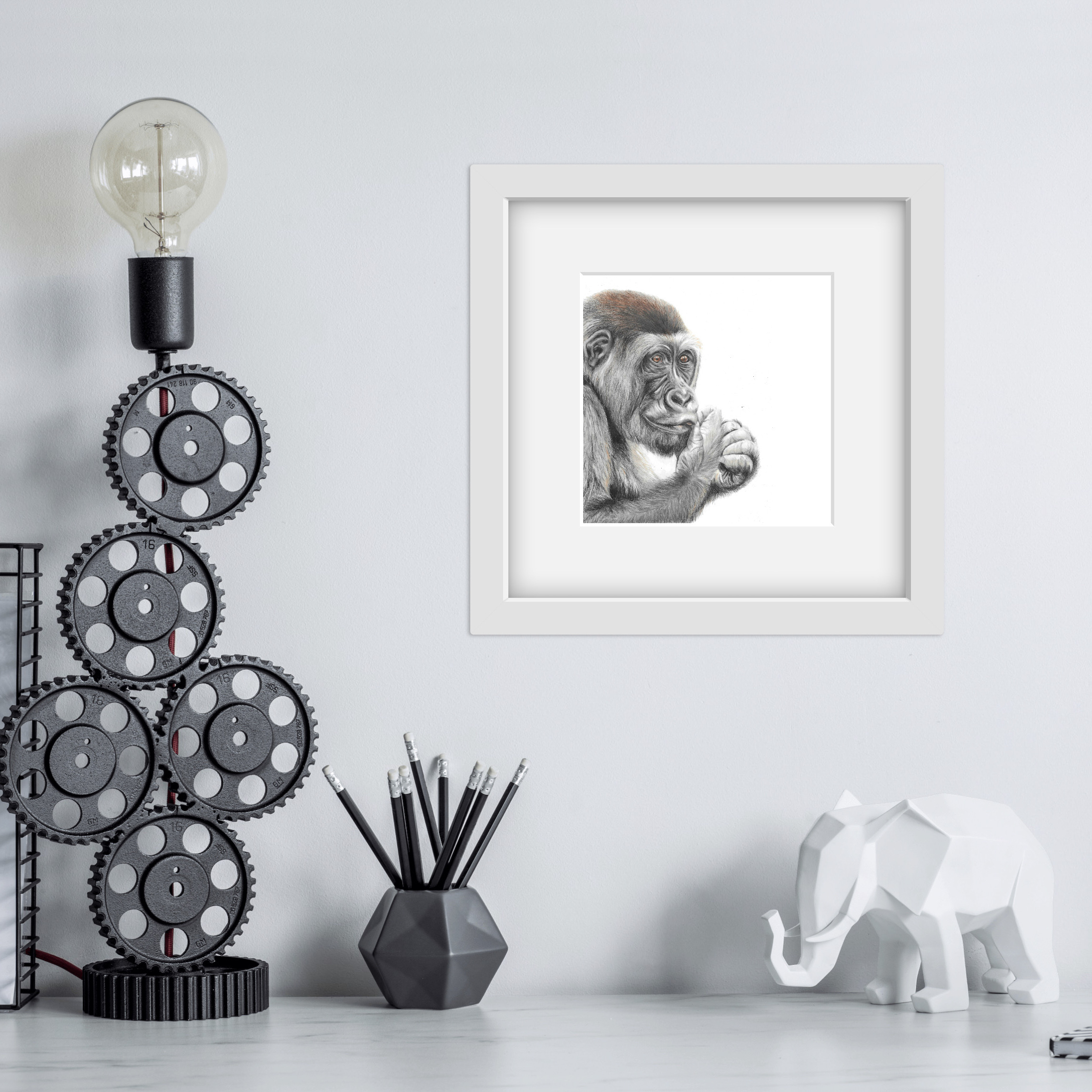 gorilla drawing in a square white frame above a shelf with black cog lamp stand and elephant model