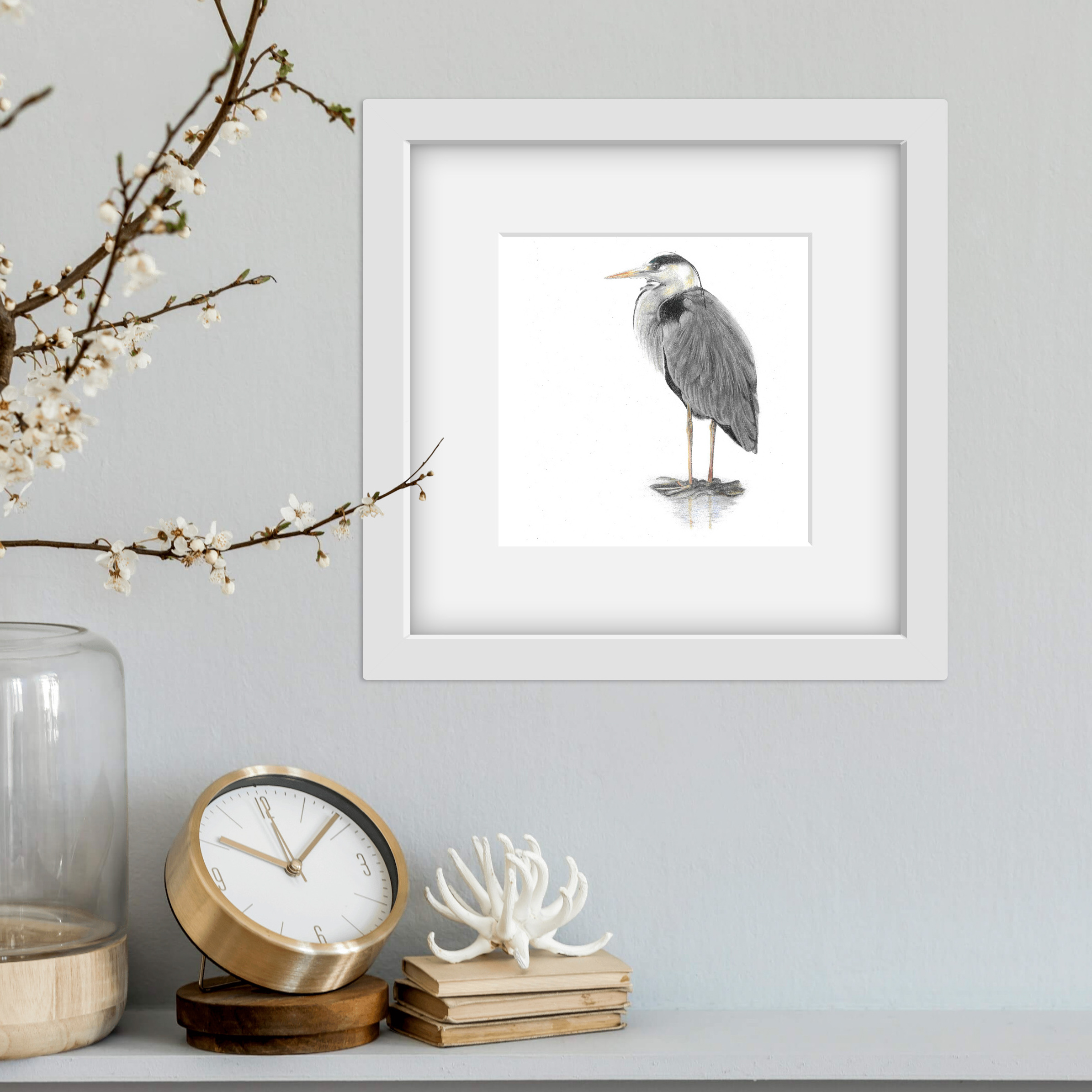 Grey heron drawing in a square white frame on a wall with clock on a shelf below